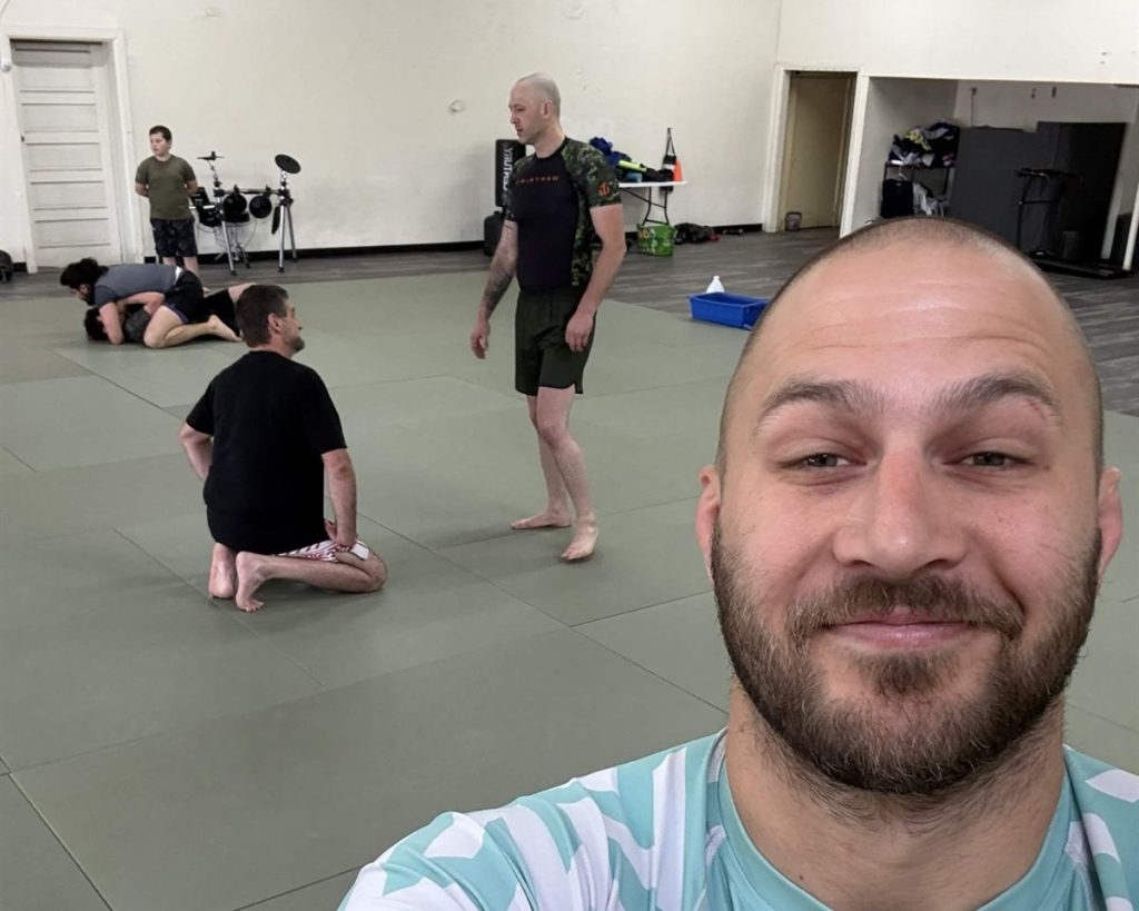 Coach Jacob smiling while students train on the mats behind him