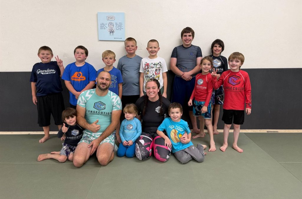 Coach Jacob and a group of kids students smiling on the mats at Consecution Jiu-Jitsu